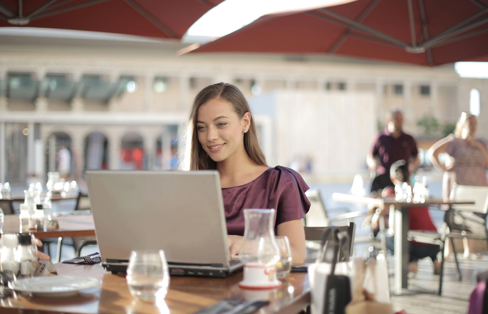 Woman in purple shirt at outdoor café with laptop, smiling on a sunny day